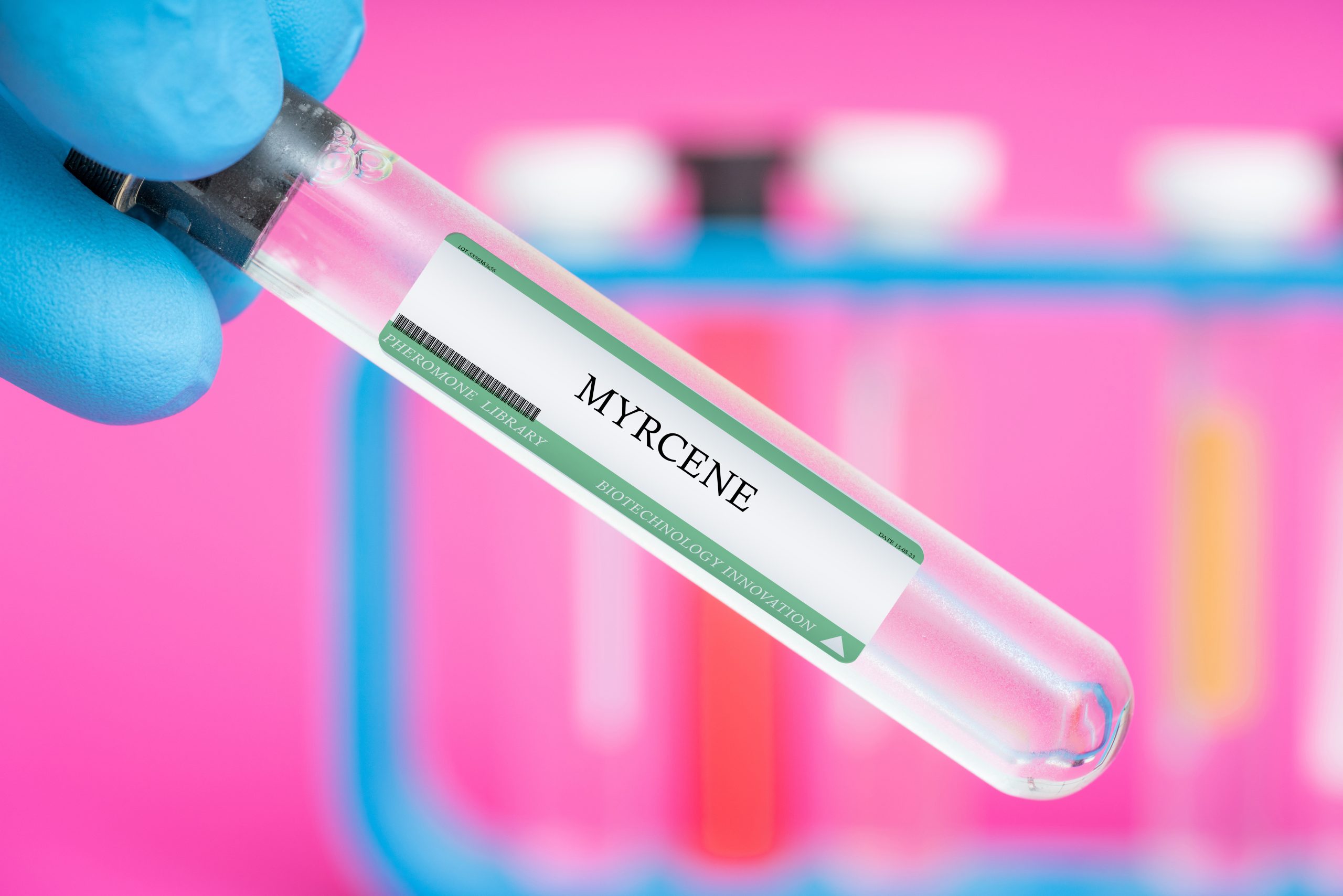 A hand holding a clear test tube that says, "Myrcene." The blurred background is pink and has other test tubes.